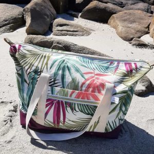 Sac week-end Sierra fermé, motif tropical et base violette, posé sur le sable face aux rochers et à la mer.
