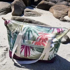 Sac week-end Sierra fermé, motif tropical et base violette, posé sur le sable face aux rochers et à la mer.