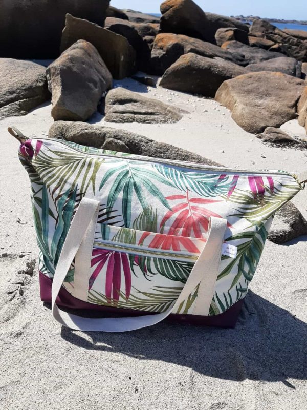 Sac week-end Sierra fermé, motif tropical et base violette, posé sur le sable face aux rochers et à la mer.