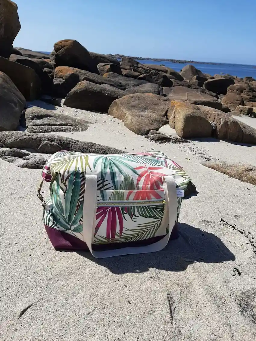 Sac week-end Sierra en version compacte, forme rectangulaire, avec mousquetons latéraux fermés. Motif tropical, base violette, posé sur le sable breton.